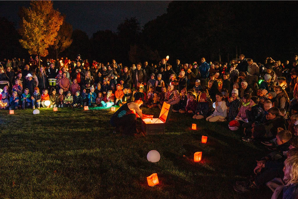 Große Menschenmenge sitzt und steht nachts im Kreis auf einer Wiese um eine Person, die eine leuchtende Truhe öffnet