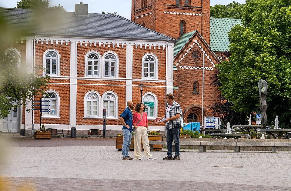 Drei Personen stehen auf einem gepflasterten Platz vor einem roten Backsteingebäude mit hohem Turm und grüner Dachspitze