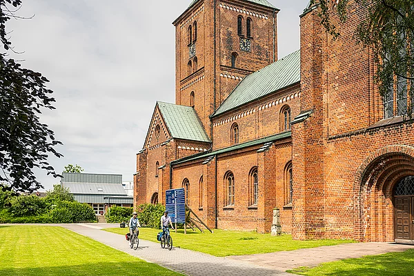 Zwei Radfahrer auf einem Weg neben einer großen Backsteinkirche mit grünem Dach und Turm