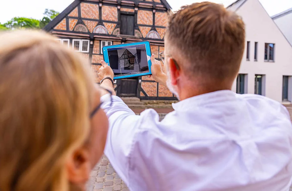 Zwei Personen betrachten gemeinsam ein Tablet mit einer historischen Gebäudedarstellung vor einem Fachwerkhaus mit offenem Tor und Museumschild