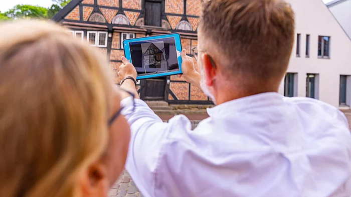 Zwei Personen betrachten gemeinsam ein Tablet mit einer historischen Gebäudedarstellung vor einem Fachwerkhaus mit offenem Tor und Museumschild