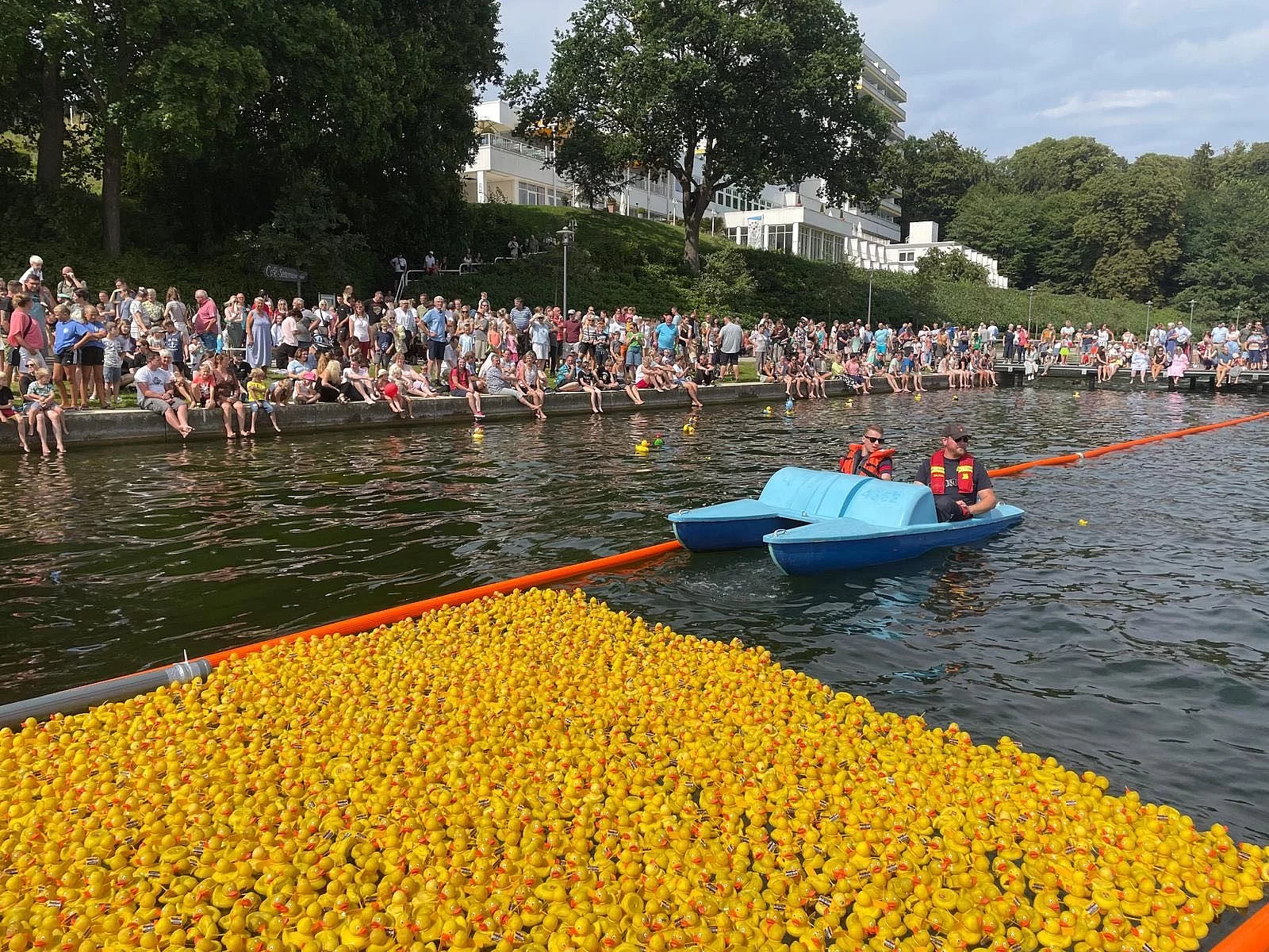 Viele gelbe Gummienten auf Wasserfläche vor Ufer mit zahlreichen Zuschauern und blauem Tretboot mit zwei Personen in Rettungswesten