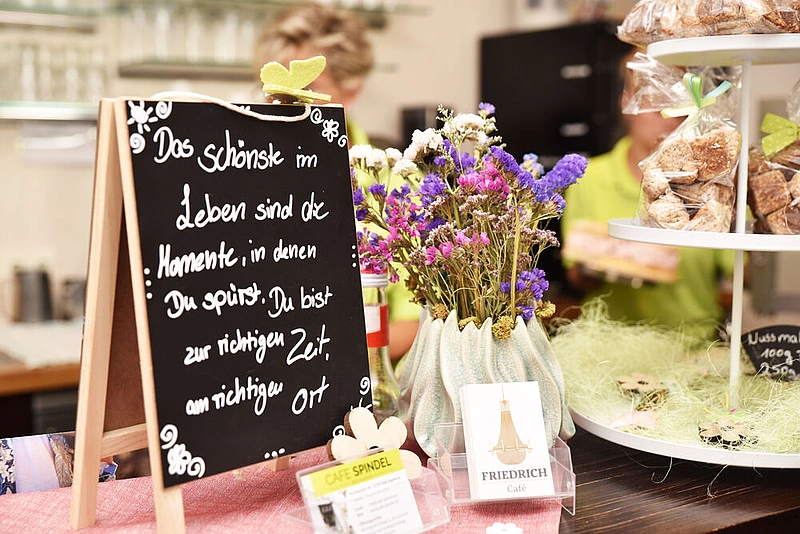 Schief stehende Tafel mit handgeschriebenem Spruch neben Vase mit lila und weißen Blumen und Auslage mit Gebäck in einem Café