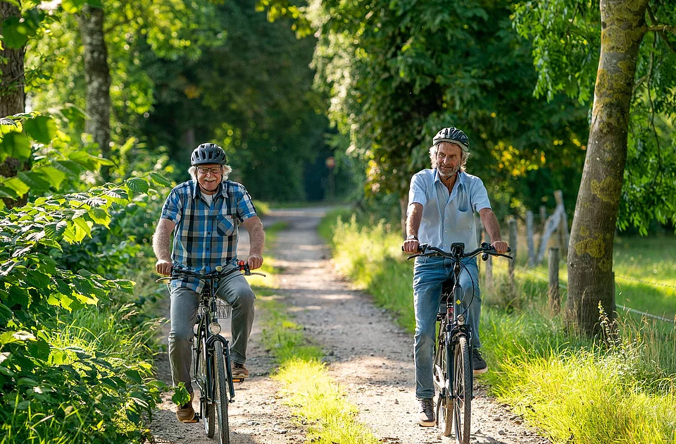Zwei Männer mit Fahrradhelmen fahren auf einem schmalen Waldweg umgeben von Bäumen und Wiesen