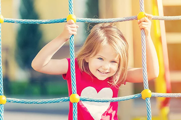 Kind mit rotem T-Shirt klettert an blau-gelben Seilnetz auf Spielplatz