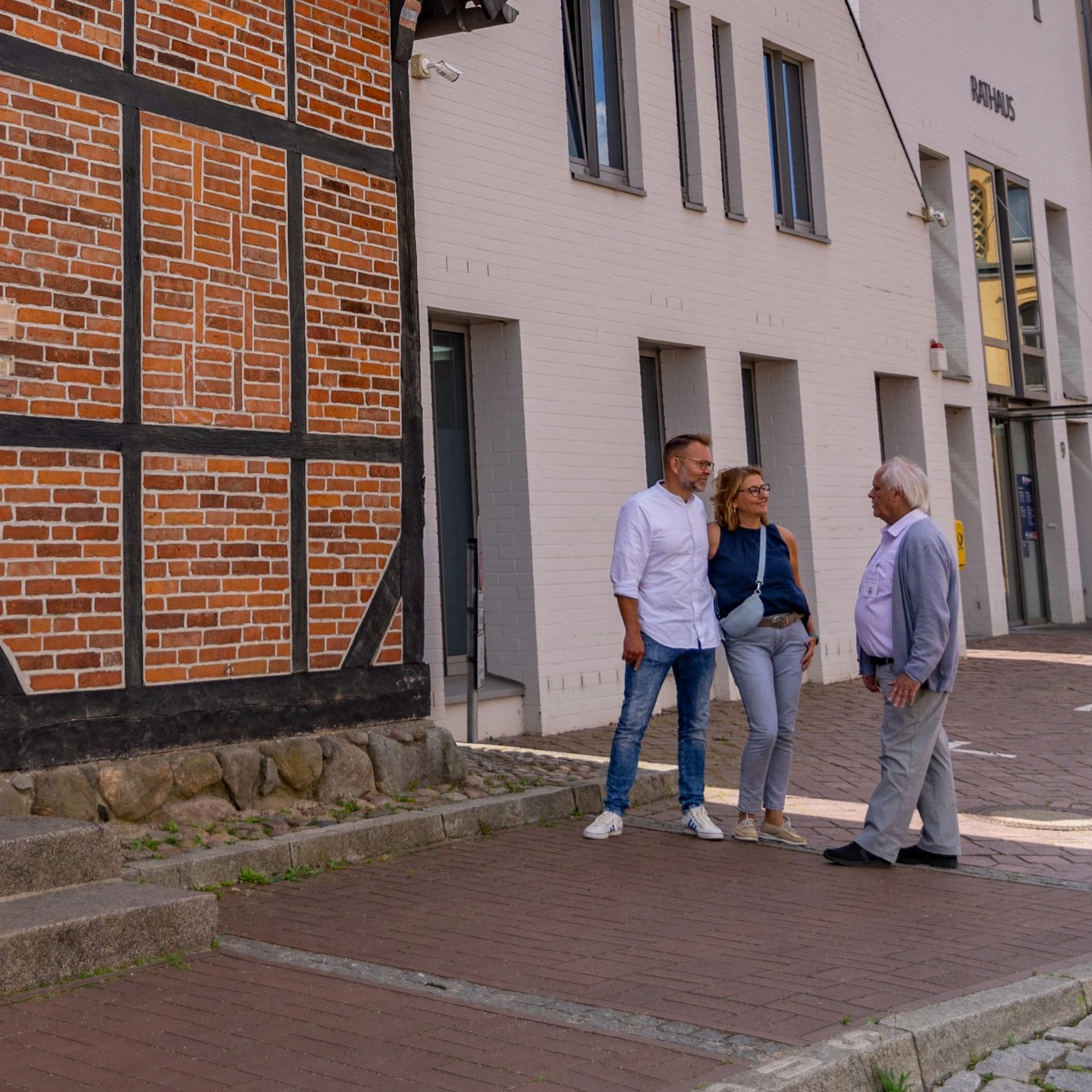 Fachwerkhaus mit rotem Backstein und schwarzem Holzfachwerk neben modernen weißen Gebäuden, davor drei Personen im Gespräch auf gepflastertem Gehweg