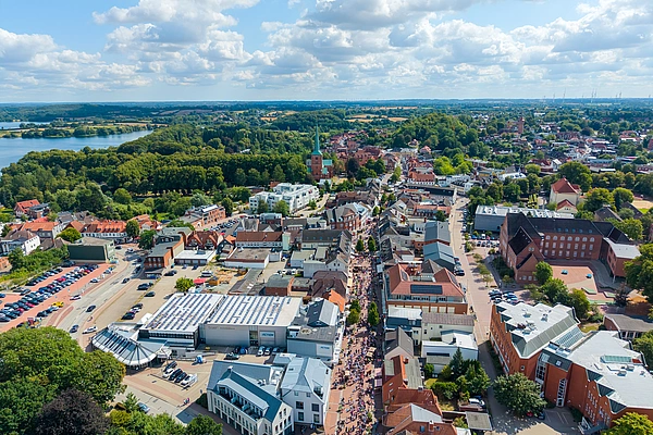 Luftaufnahme einer Stadt, einem See im Hintergrund und bewaldeten Flächen unter blauem Himmel mit Wolken
