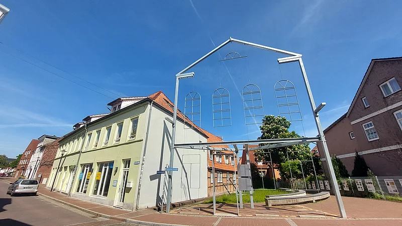 Metallrahmen in Form der Fassade einer ehemaligen Synagoge vor einem hellgrünen Gebäude in einer ruhigen Straße bei blauem Himmel