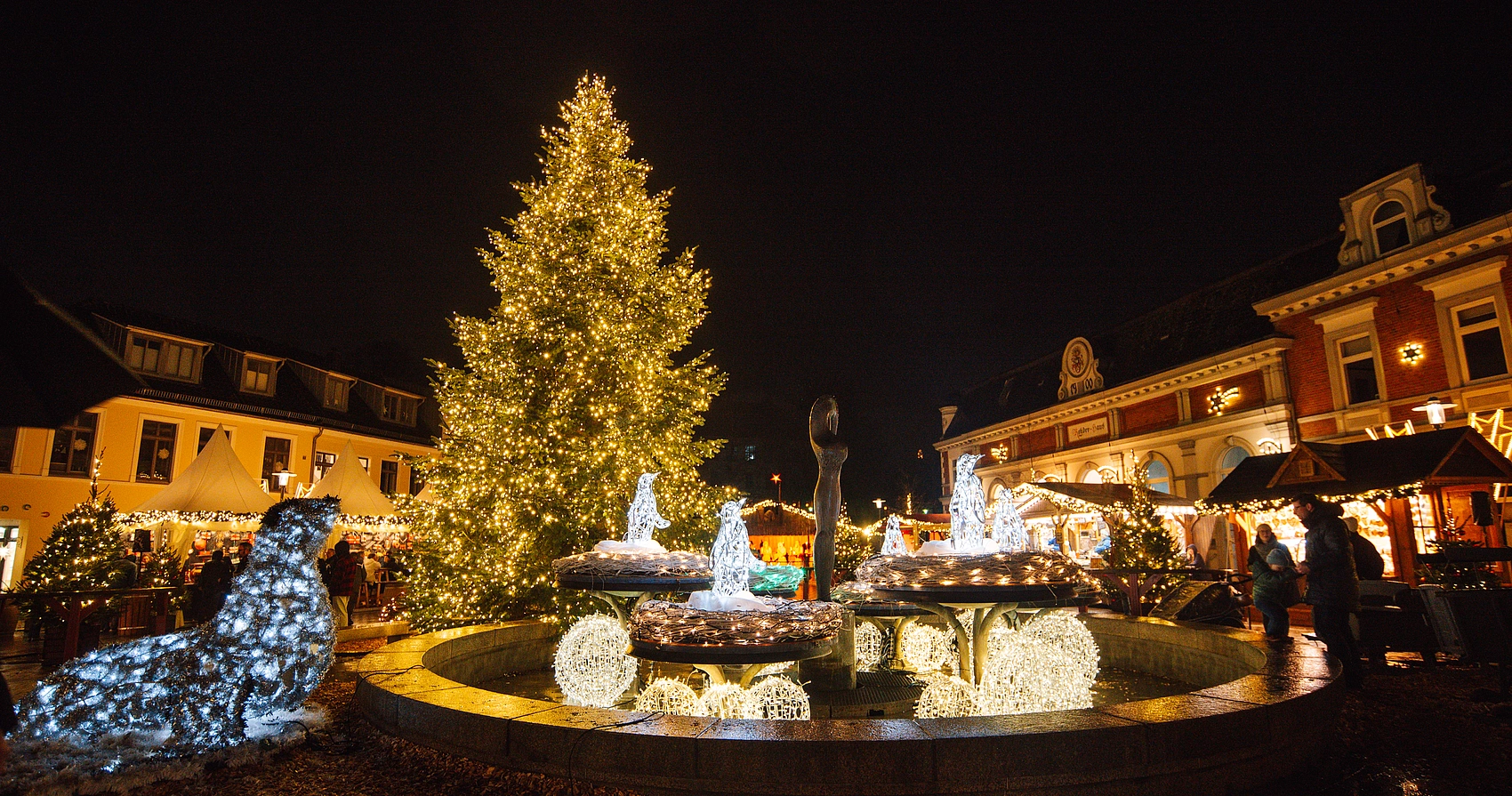 Weihnachtsmarkt bei Nacht mit großem beleuchtetem Weihnachtsbaum, beleuchteten Tierfiguren und einem Brunnen im Vordergrund, umgeben von historischen Gebäuden und beleuchteten Holzbuden