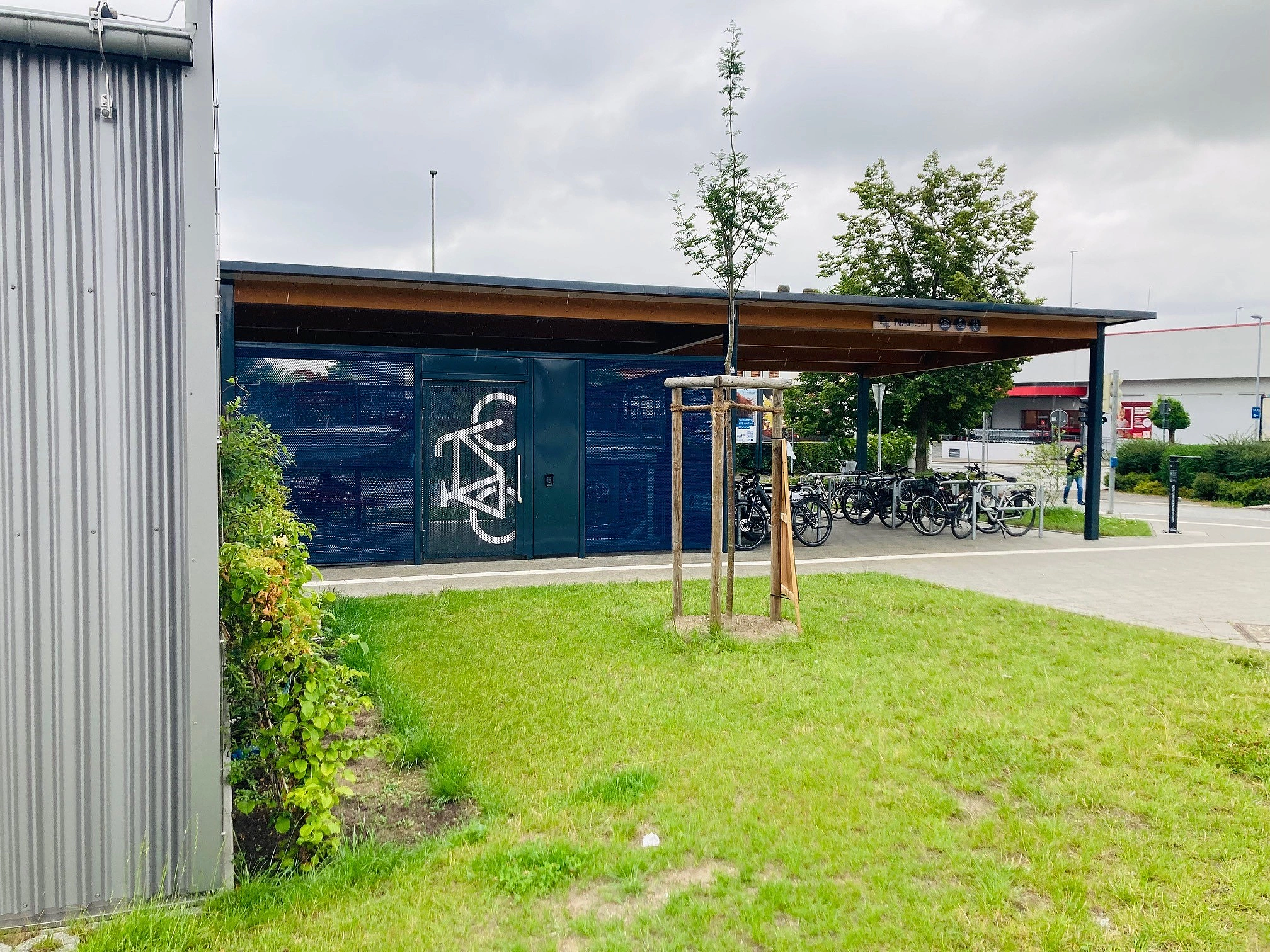 Fahrradunterstand mit Holzdach und blauer Wand mit weißem Fahrradsymbol neben einem kleinen Baum auf einer Rasenfläche
