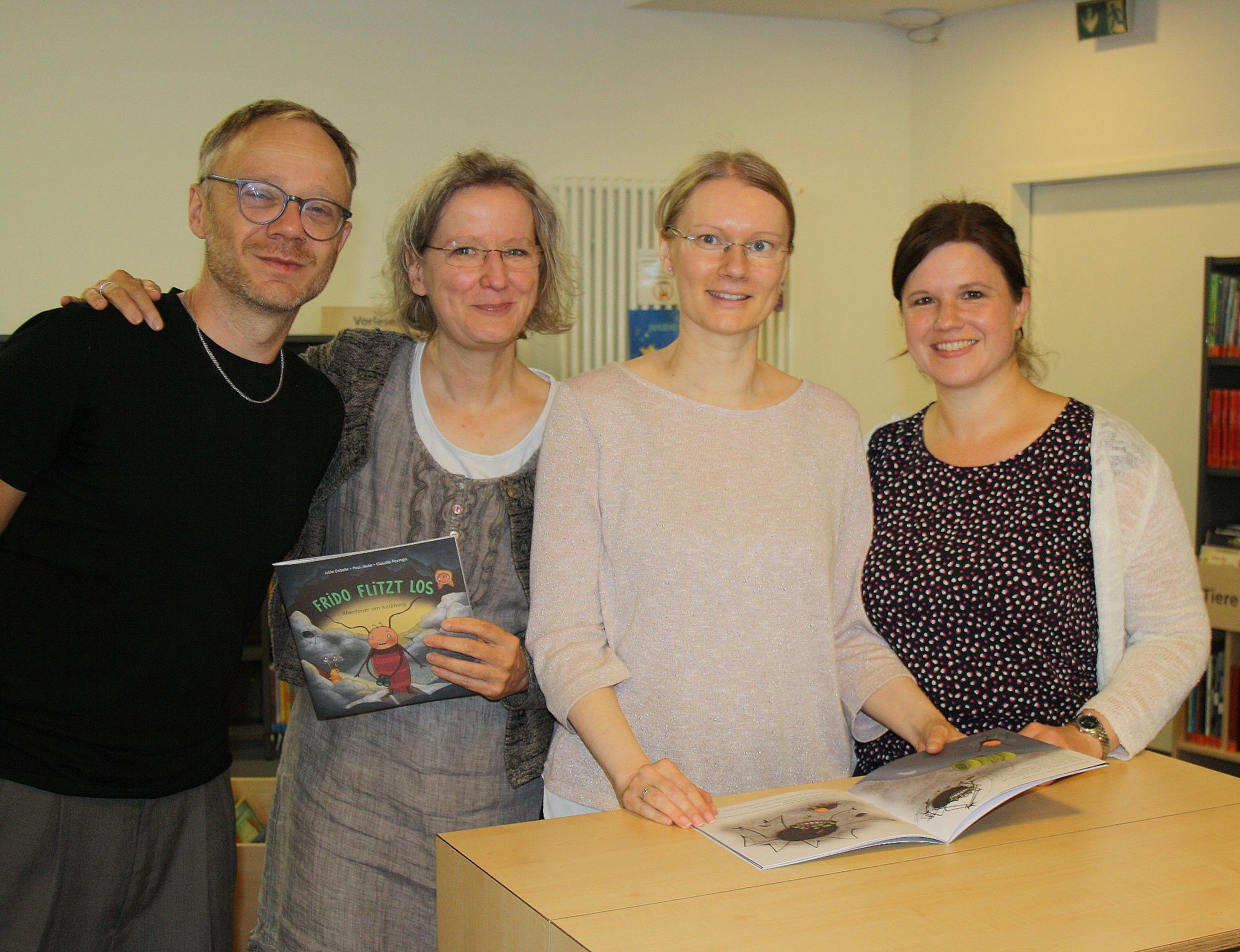 Vier lächelnde Personen stehen an einem Tisch, eine hält ein Buch mit farbiger Illustration, eine andere ein aufgeschlagenes Buch mit Tierzeichnungen