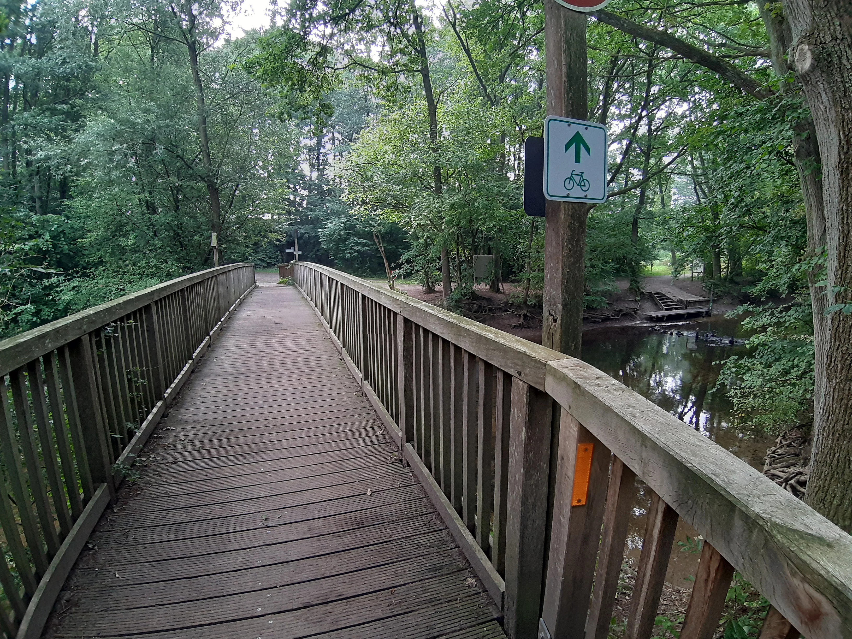 Holzbrücke mit Geländer über einen Fluss in einem bewaldeten Gebiet mit Fahrradweg-Schild