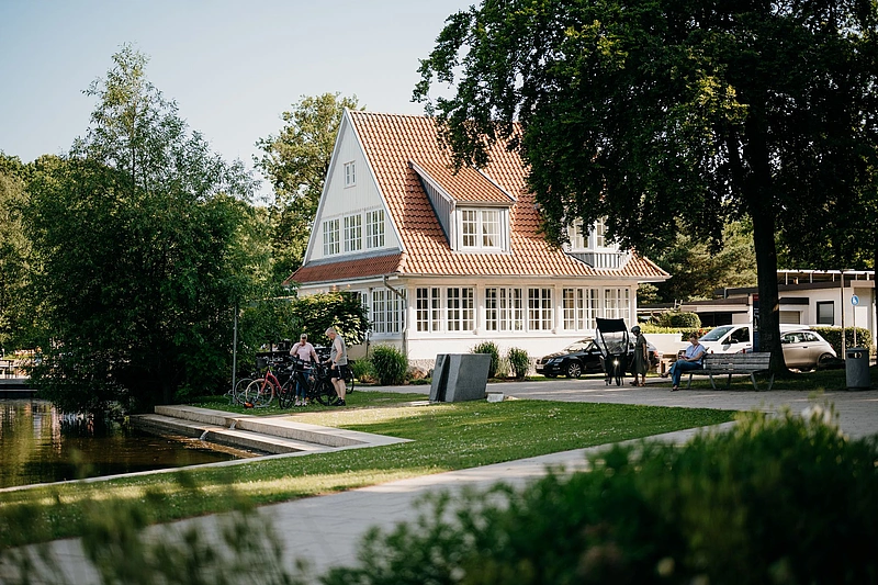 Zweistöckiges Haus mit Satteldach und vielen Fenstern an einem Seeufer mit Menschen, Fahrrädern und parkenden Autos