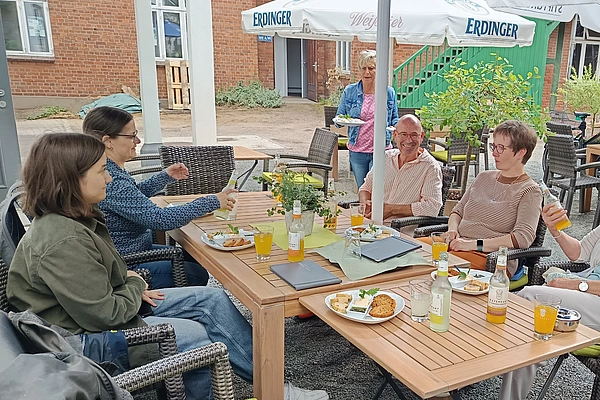 Gruppe von fünf Personen sitzt an zwei zusammengestellten Holztischen im Außenbereich eines Cafés und trinkt Getränke