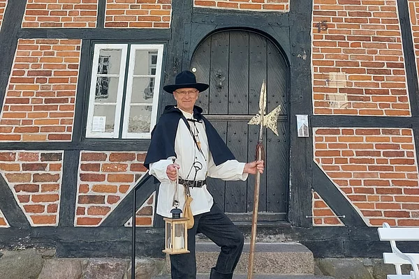 Person in historischer Nachtwächterkleidung mit Laterne und Hellebarde steht vor einem Fachwerkhaus mit Ziermauerwerk und einer Tür mit Rundbogen