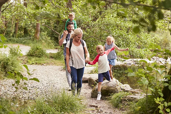 Familie mit zwei Kindern und einem Kleinkind auf dem Rücken des Vaters wandert auf einem Waldweg neben einem See