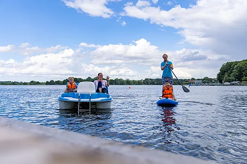Zwei Kinder mit orangefarbenen Schwimmwesten auf einem Tretboot und einem Stand-up-Paddle-Board auf einem See bei bewölktem Himmel