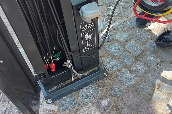 Fahrradpumpe mit Manometer und Symbolen für Fahrrad, Rollstuhl und Kinderwagen auf gepflastertem Boden, daneben Hände, die an einem Fahrradreifen arbeiten