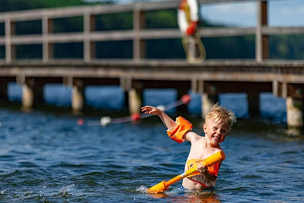 Kind mit orangefarbenen Schwimmflügeln spielt mit einem orangefarbenen Spielzeug im Wasser vor einem Steg am Großen Segeberger See