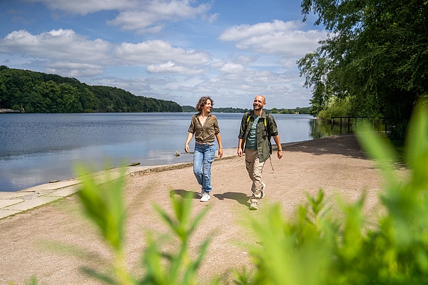 Zwei Personen gehen an einem Seeuferweg bei sonnigem Wetter, im Vordergrund unscharfe grüne Blätter