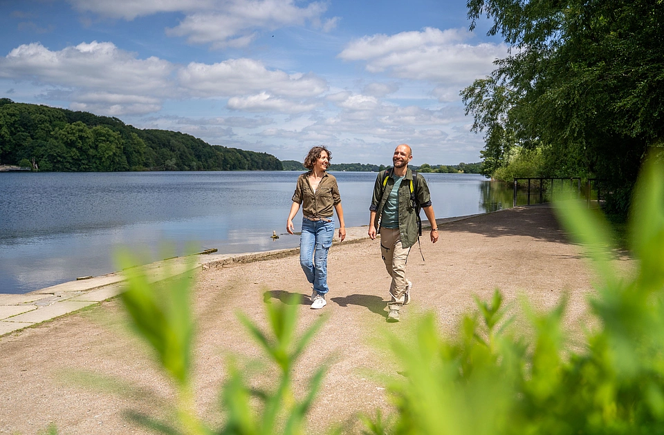 Zwei Personen gehen an einem Seeuferweg bei sonnigem Wetter, im Vordergrund unscharfe grüne Blätter
