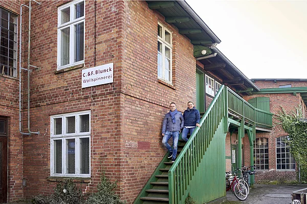 Zwei Personen stehen auf einer grünen Außentreppe an einem roten Backsteingebäude mit dem Schild 'C. & F. Blunck Wollspinnerei'