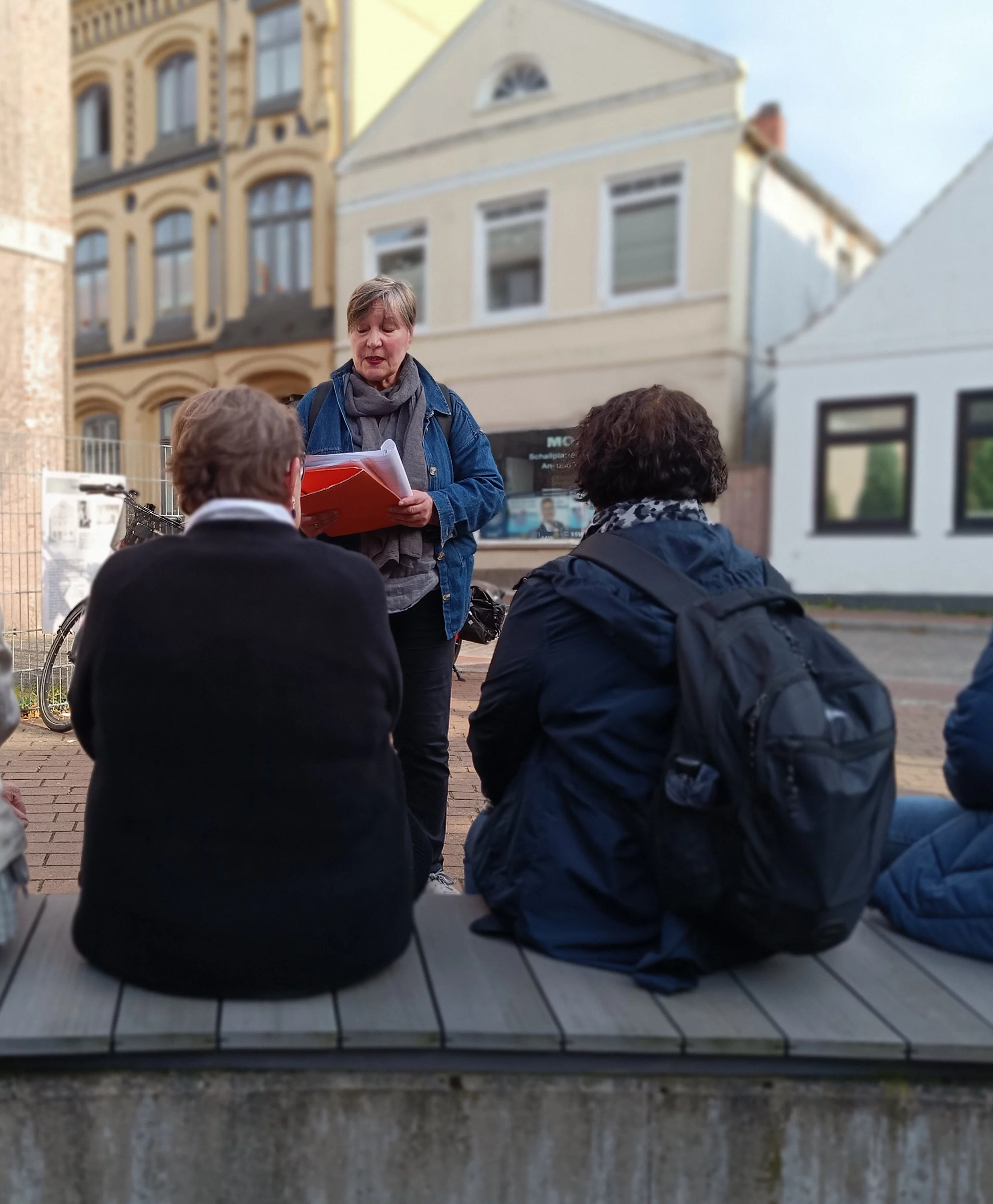 Drei Frauen bei einem Spaziergang sitzen auf einer Bank in einer Stadt, eine steht und hält Papiere in der Hand, im Hintergrund Häuserfassaden.