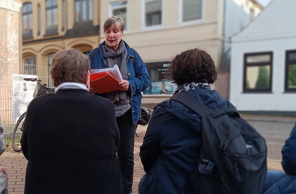 Drei Frauen bei einem Spaziergang sitzen auf einer Bank in einer Stadt, eine steht und hält Papiere in der Hand, im Hintergrund Häuserfassaden.