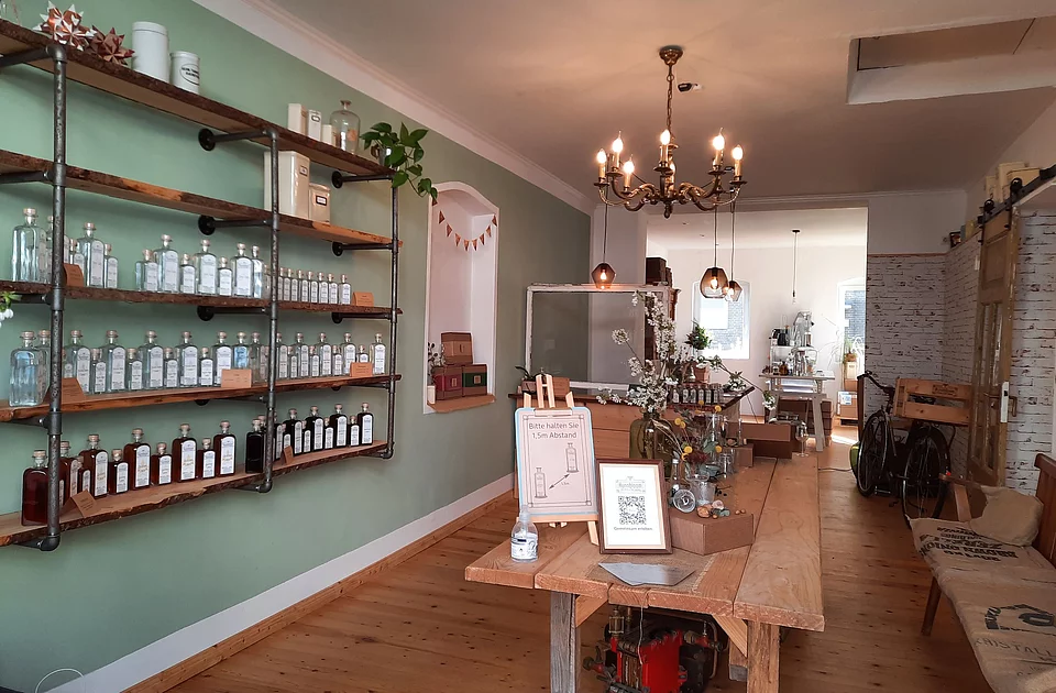 Innenraum eines Ladens mit Holzboden, Regalen voller Flaschen an der Wand, einem langen Holztisch mit Dekoration und Sitzbank mit Kissen