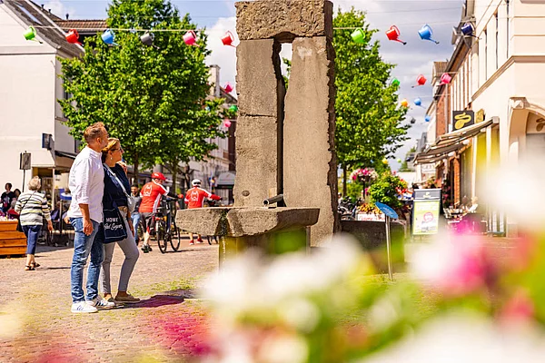Janusbrunnen aus Stein in der Fußgängerzone von Bad Segeberg mit zwei Personen, die ihn betrachten, und bunten Blumentöpfen im Vordergrund