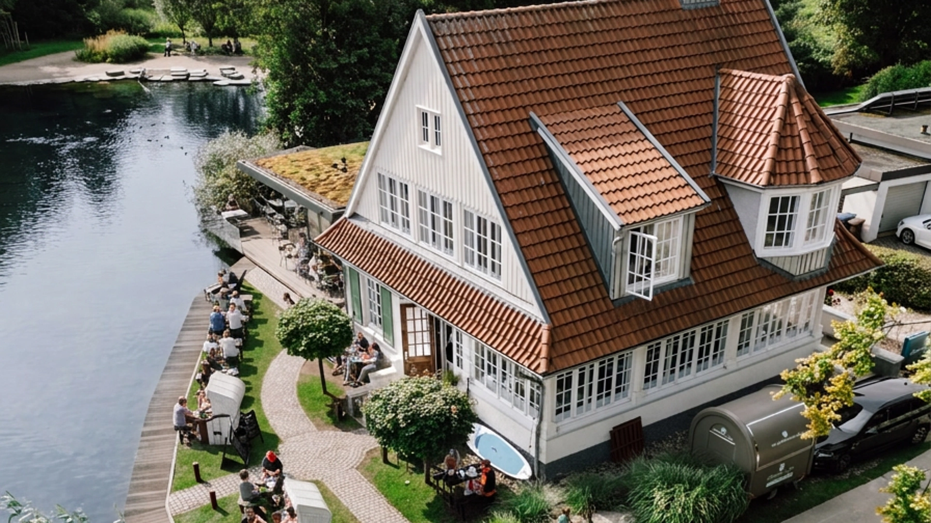 Luftaufnahme eines weißen Hauses mit rotem Dach am Seeufer, davor sitzen Menschen an Tischen auf einer Terrasse