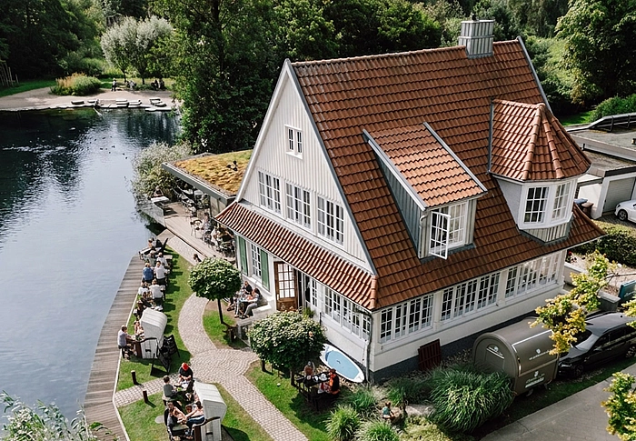 Luftaufnahme eines weißen Hauses mit rotem Dach am Seeufer, davor sitzen Menschen an Tischen auf einer Terrasse
