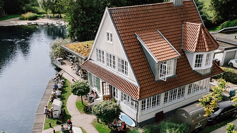 Goldmarie am See Luftaufnahme eines weißen Hauses mit rotem Dach am Seeufer, davor sitzen Menschen an Tischen auf einer Terrasse