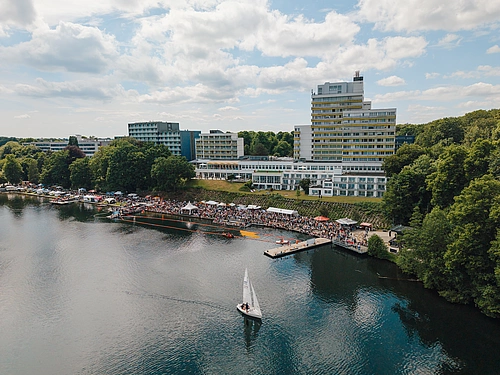 Luftaufnahme eines Seefests mit Segelboot auf dem Wasser, Menschenansammlung am Ufer und mehrstöckigen Gebäuden im Hintergrund