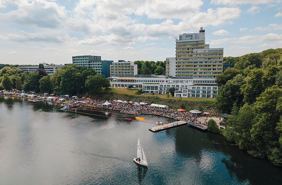 Luftaufnahme eines Seefests mit Segelboot auf dem Wasser, Menschenansammlung am Ufer und mehrstöckigen Gebäuden im Hintergrund