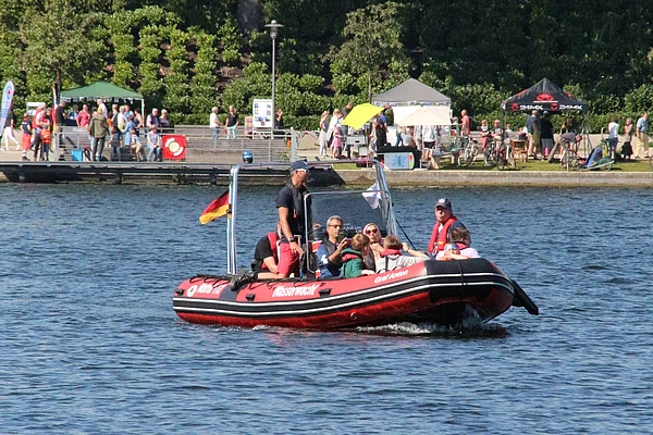 Mehrere Personen sitzen in einem roten Schlauchboot mit deutscher Flagge auf einem See vor einer Uferpromenade mit Menschen, Zelten und Bäumen beim Bad Segeberger Seefest.