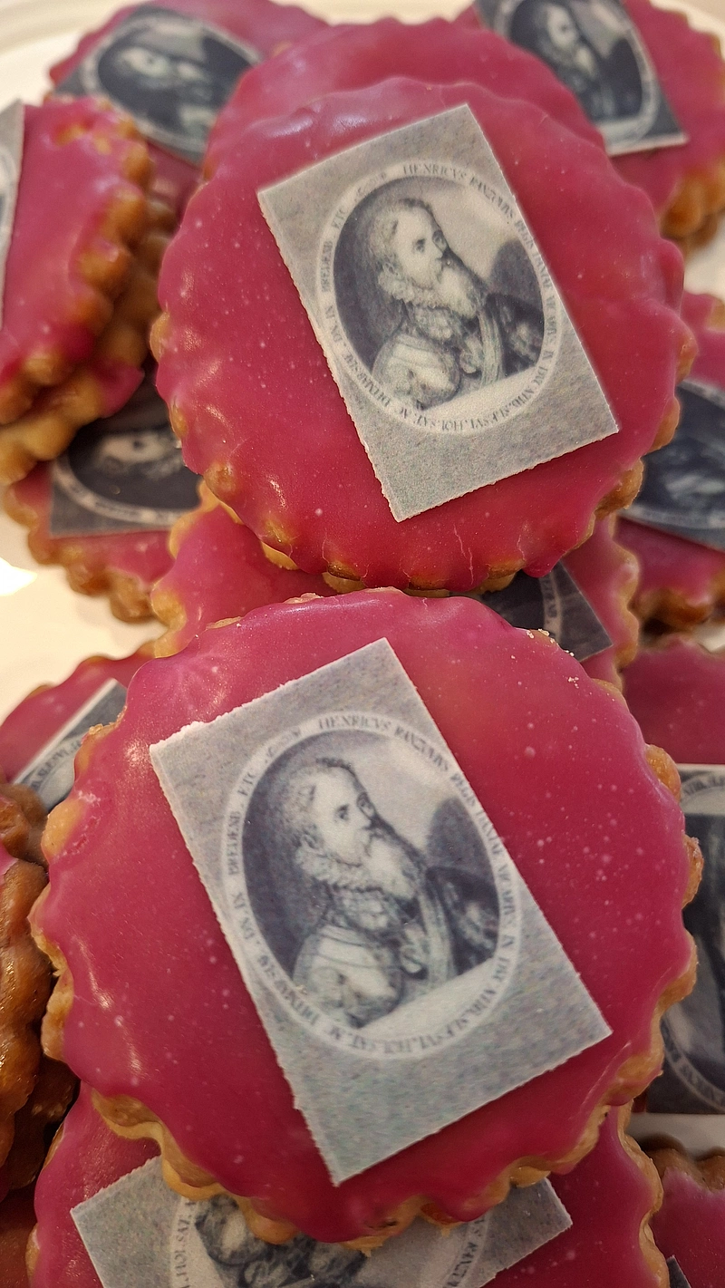 Mehrere runde, gezackte Kekse mit rotem Zuckerguss und einem kleinen schwarz-weißen Porträt eines Mannes auf Papierstücken darauf