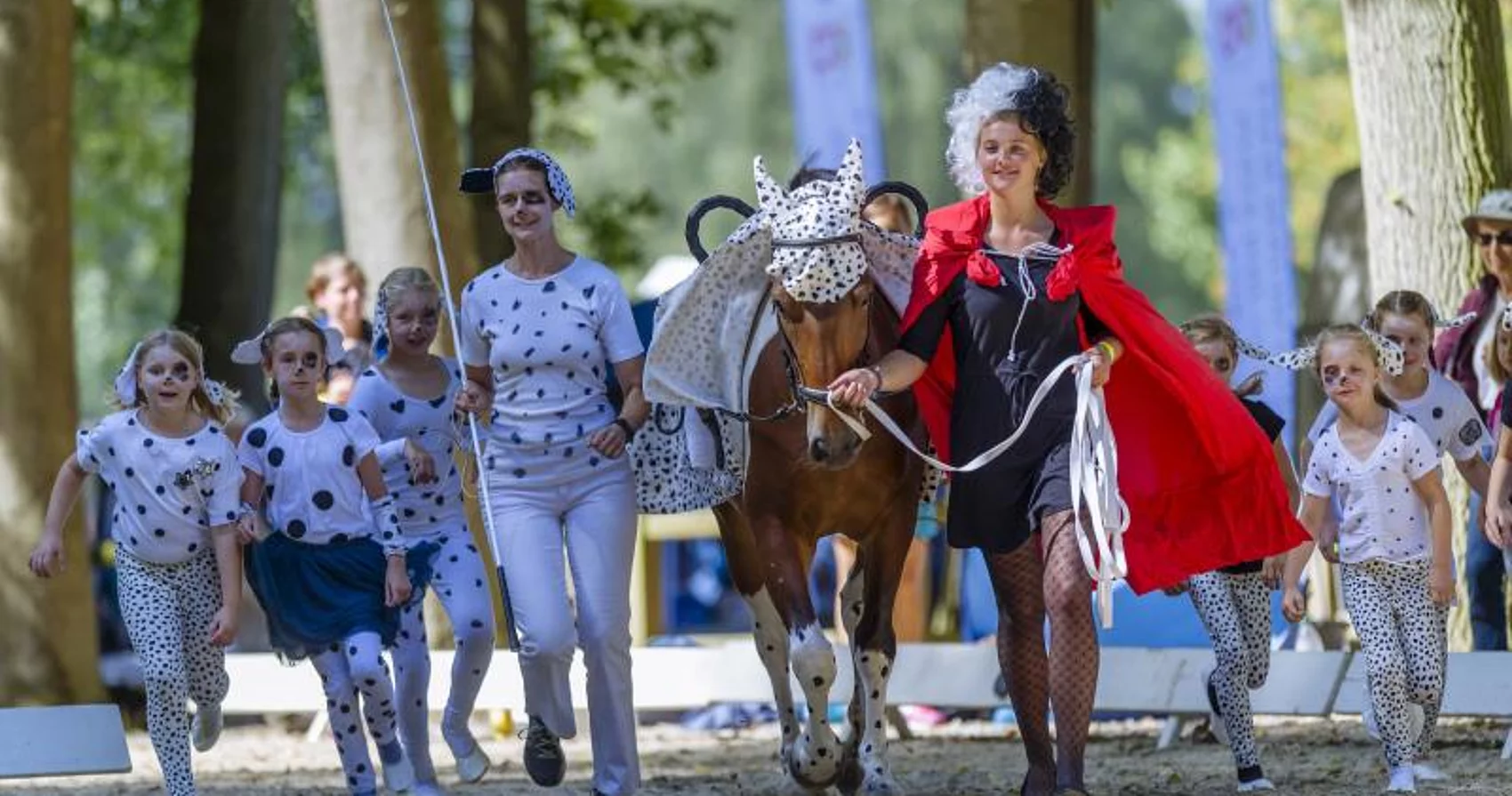 Gruppe von Kindern und zwei Erwachsenen in gepunkteten Kostümen führen ein geschecktes Pferd, eine Frau trägt ein rotes Cape und schwarze Netzstrumpfhosen