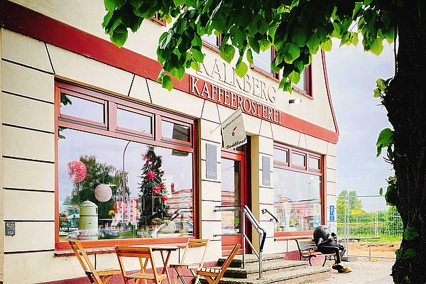 Außenansicht einer Kaffeerösterei mit Tischen und Stühlen vor dem Eingang und einer Person auf einer Bank neben dem Gebäude