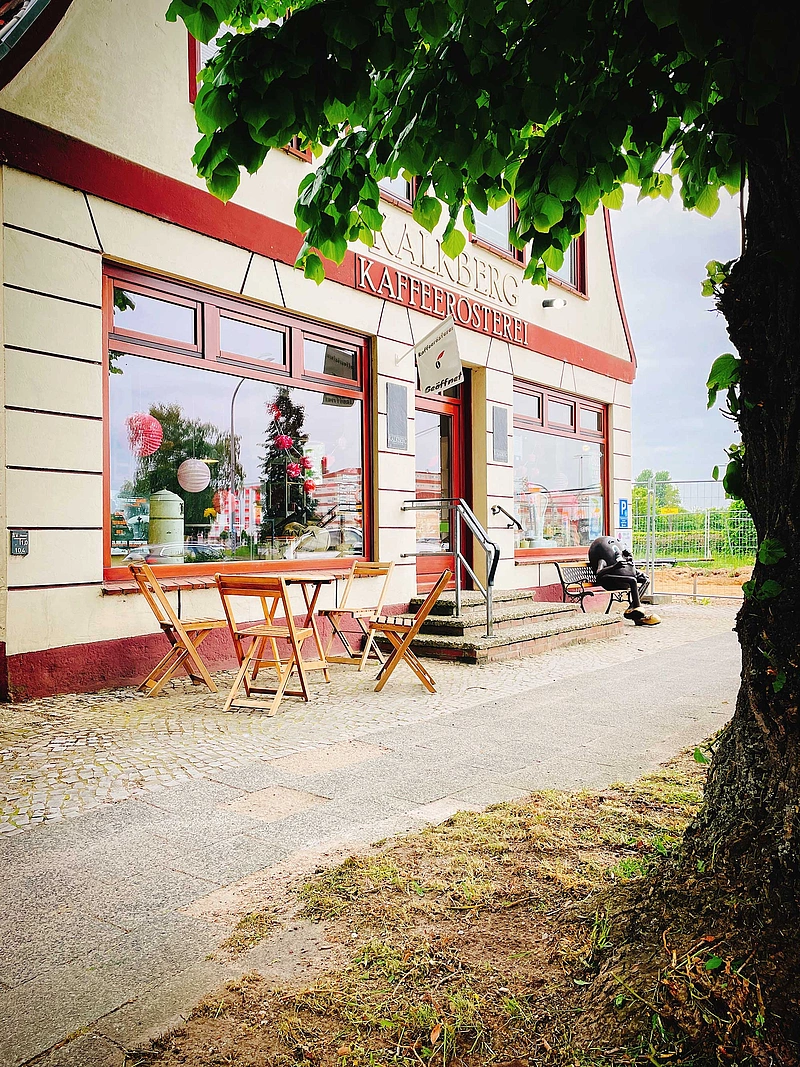 Außenansicht einer Kaffeerösterei mit Tischen und Stühlen vor dem Eingang und einer Person auf einer Bank neben dem Gebäude