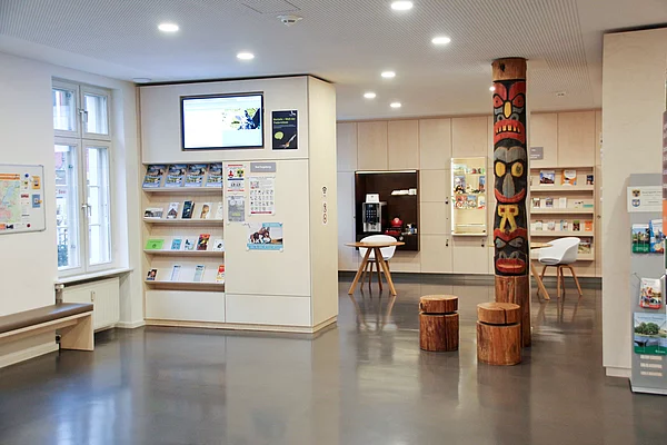 Innenraum einer Tourist-Information mit Sitzbank, Informationsregalen, einem bemalten Totempfahl und Sitzgelegenheiten aus Baumstämmen