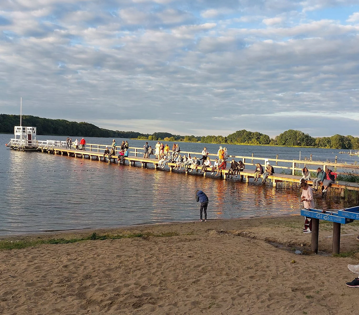 Menschen sitzen und stehen auf einem langen Steg über einem See bei bewölktem Himmel