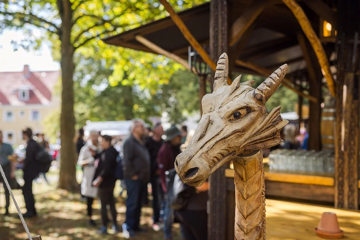 Nahaufnahme einer hölzernen Drachenkopfschnitzerei vor einem Marktstand mit Menschen im Hintergrund