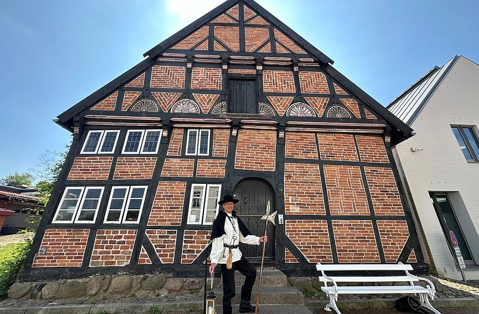 Person in historischer Kleidung mit Hut, Umhang, Stiefeln, Laterne in der linken Hand und Speer in der rechten Hand vor Fachwerkhaus mit Holztür