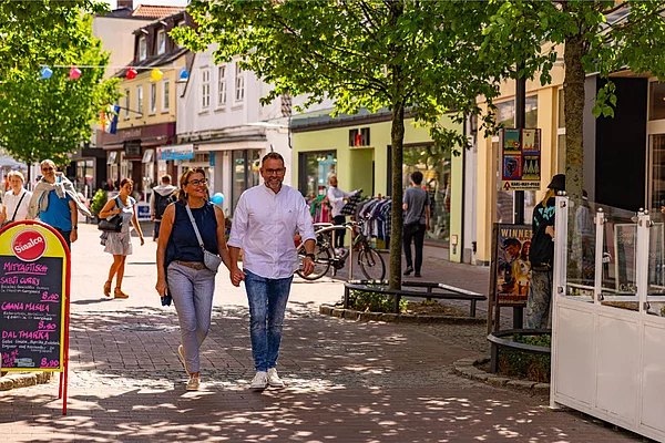 Paar hält Händchen und geht auf einer von Bäumen gesäumten Fußgängerzone mit Geschäften und Passanten spazieren