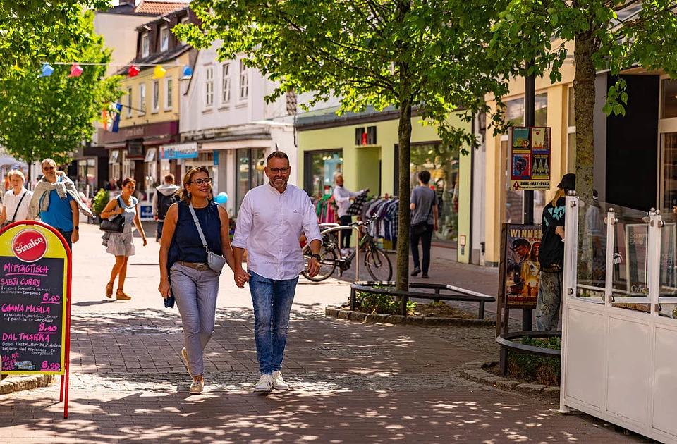 Paar hält Händchen und geht auf einer von Bäumen gesäumten Fußgängerzone mit Geschäften und Passanten spazieren