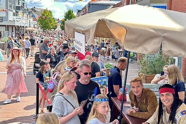 Menschen stehen in einer Schlange vor einem Stand mit zwei Männern in Kostümen, im Hintergrund eine belebte Straße mit weiteren Passanten und bunten Lampions über der Straße