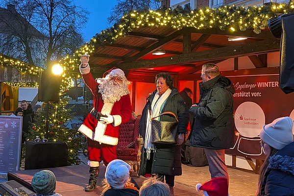 Weihnachtsmann mit erhobener Hand auf einer festlich beleuchteten Bühne vor einem Publikum mit Kindern in Weihnachtsmützen in Bad Segeberg