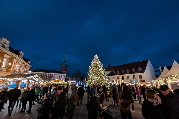 Weihnachtsmarkt bei Abenddämmerung mit beleuchtetem Weihnachtsbaum und Menschenmenge zwischen festlich geschmückten Ständen