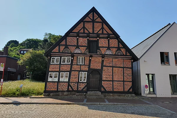 Fachwerkhaus mit Ziegelfüllung und schwarzem Holzrahmen neben modernem weißem Gebäude auf gepflastertem Platz